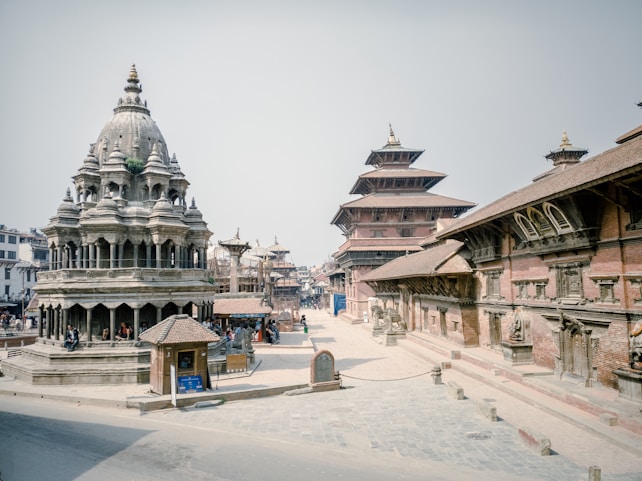 A historic architectural scene featuring traditional Nepalese pagoda-style temples and intricately carved stone structures. The setting appears to be a public square, with several people scattered throughout the area, some sitting near the temple and others walking. The temples are made of a combination of stone and wood, with unique tiered roofs that are characteristic of the region.