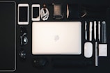 A neatly arranged collection of everyday items is displayed on a dark surface. At the center is an open laptop, surrounded by various objects such as two smartphones, a set of earphones, multiple pens, a mouse, sunglasses, a watch, a wallet, a small notebook, a ruler, a packet of mints, and a metal pen. The arrangement suggests organization and preparedness.