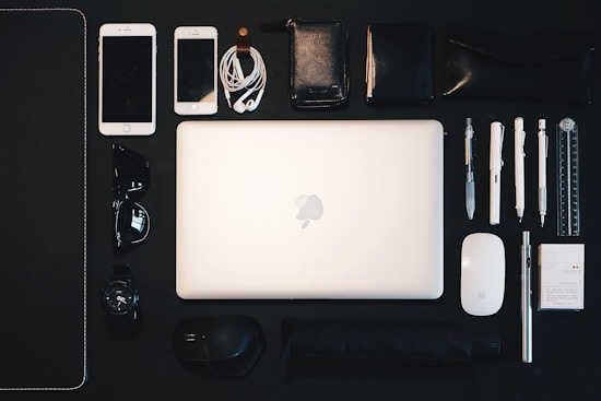 A neatly arranged collection of everyday items is displayed on a dark surface. At the center is an open laptop, surrounded by various objects such as two smartphones, a set of earphones, multiple pens, a mouse, sunglasses, a watch, a wallet, a small notebook, a ruler, a packet of mints, and a metal pen. The arrangement suggests organization and preparedness.