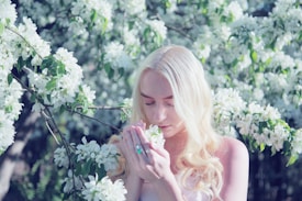 A person with long blonde hair stands surrounded by branches of white flowers. They are gently holding a cluster of blossoms close to their face, eyes closed, conveying a sense of serenity and connection with nature.