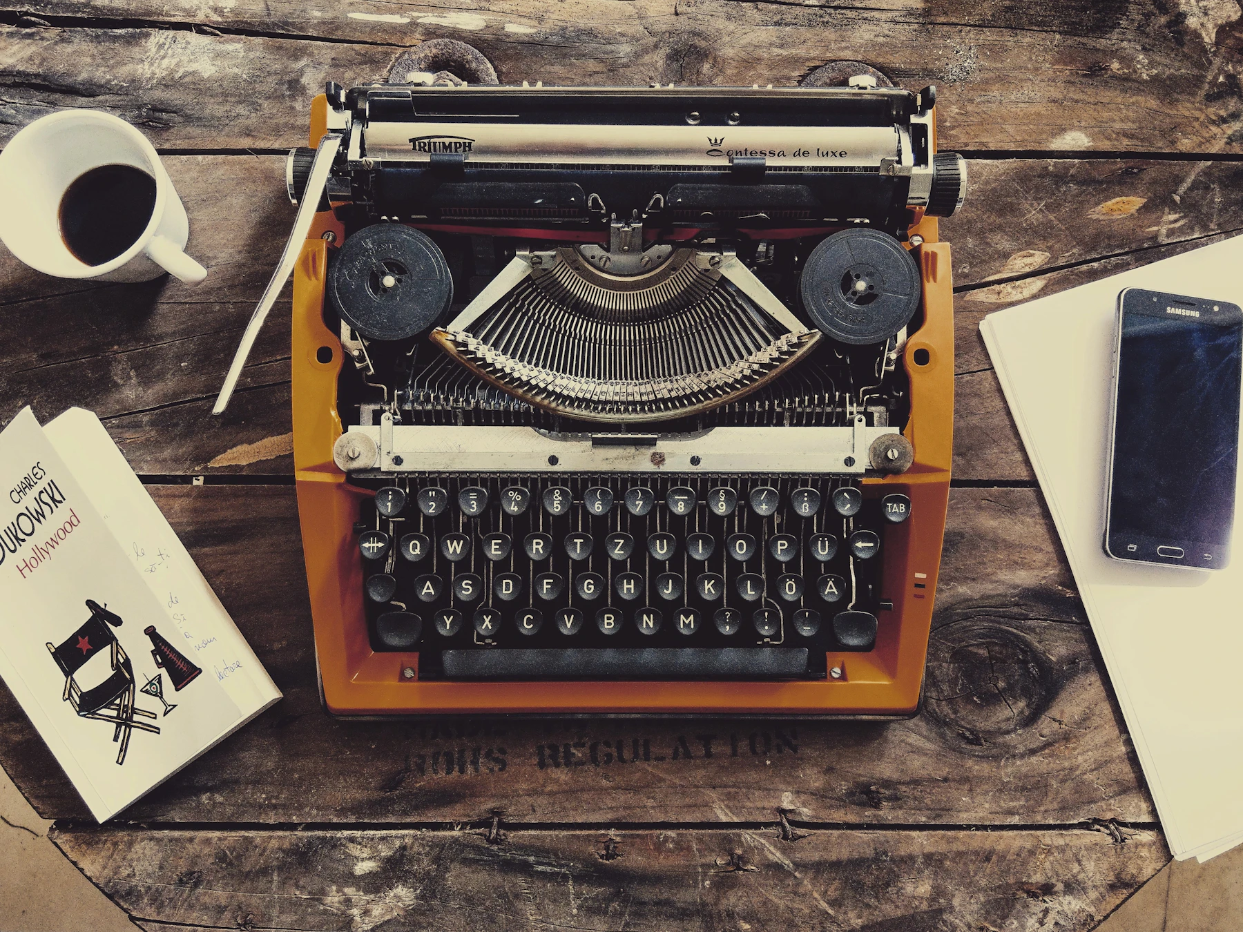 An orange typewriter on a wooden desk beside a cup of coffee, a book and a phone
