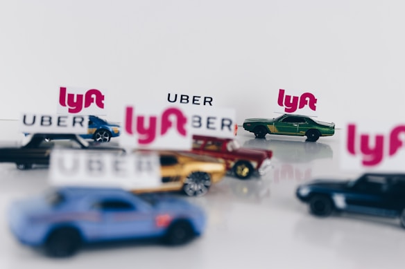 Several miniature toy cars are arranged with blurred Uber and Lyft logos. The cars are differently colored, including shades of blue, green, and gold, and are set against a plain background.