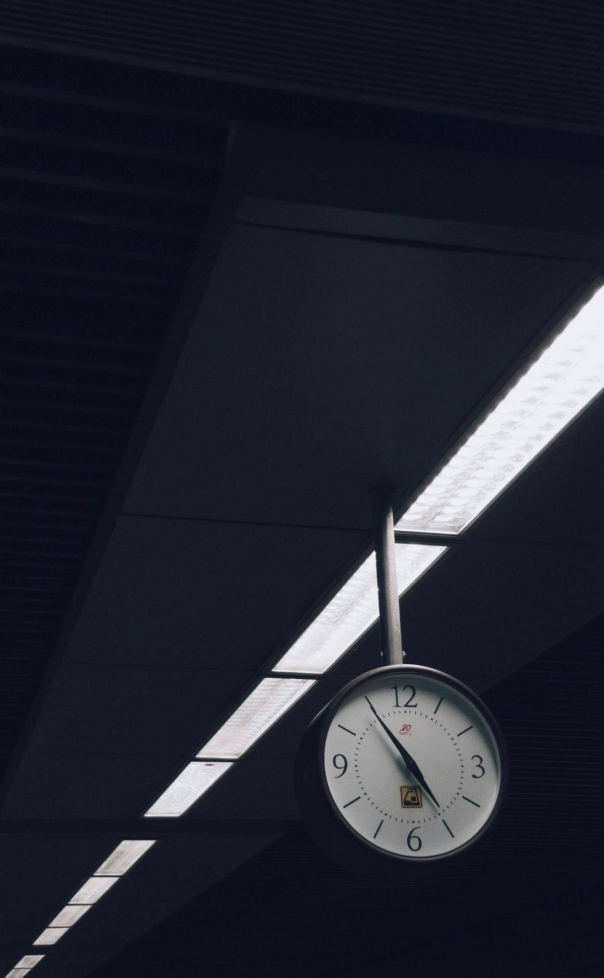 A station-style analog clock suspended from a ceiling beneath skylight strips, shared public time
