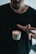 A person is wearing a dark t-shirt and appears to be holding a small cup of coffee with latte art. The coffee cup is shown mid-air, giving an illusion of levitation as the person's fingers are positioned as if about to grasp it.