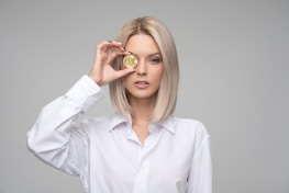 A person with blond hair wearing a white shirt holds a gold coin embossed with a cryptocurrency symbol in front of one eye. The background is plain and gray, focusing attention on the subject.
