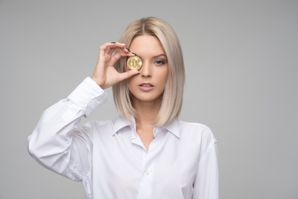 A person with blond hair wearing a white shirt holds a gold coin embossed with a cryptocurrency symbol in front of one eye. The background is plain and gray, focusing attention on the subject.