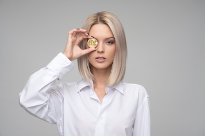 A person with blond hair wearing a white shirt holds a gold coin embossed with a cryptocurrency symbol in front of one eye. The background is plain and gray, focusing attention on the subject.