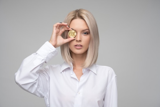 A person with blond hair wearing a white shirt holds a gold coin embossed with a cryptocurrency symbol in front of one eye. The background is plain and gray, focusing attention on the subject.