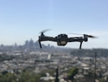 A drone with four rotors is hovering in mid-air against a backdrop of a blurred city skyline. The propellers are in motion, giving an impression of flight. The cityscape below is out of focus, enhancing the prominence of the drone.