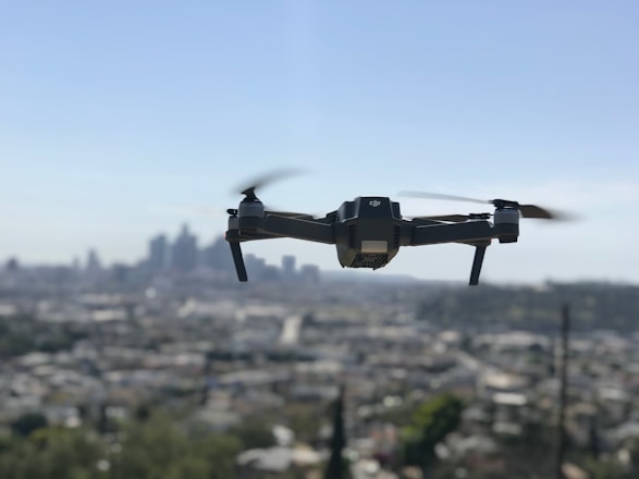 A drone with four rotors is hovering in mid-air against a backdrop of a blurred city skyline. The propellers are in motion, giving an impression of flight. The cityscape below is out of focus, enhancing the prominence of the drone.