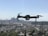 A drone with four rotors is hovering in mid-air against a backdrop of a blurred city skyline. The propellers are in motion, giving an impression of flight. The cityscape below is out of focus, enhancing the prominence of the drone.