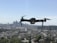 A drone with four rotors is hovering in mid-air against a backdrop of a blurred city skyline. The propellers are in motion, giving an impression of flight. The cityscape below is out of focus, enhancing the prominence of the drone.