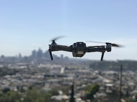 A drone with four rotors is hovering in mid-air against a backdrop of a blurred city skyline. The propellers are in motion, giving an impression of flight. The cityscape below is out of focus, enhancing the prominence of the drone.