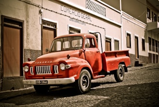 A vintage red Bedford truck is parked on a cobblestone street in front of a building with a sign reading 'Comercio Geral'. The building has a simple facade with several doors and windows. The scene has a nostalgic and historical feel.