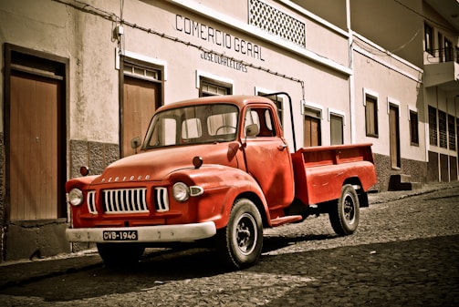 A vintage red Bedford truck is parked on a cobblestone street in front of a building with a sign reading 'Comercio Geral'. The building has a simple facade with several doors and windows. The scene has a nostalgic and historical feel.