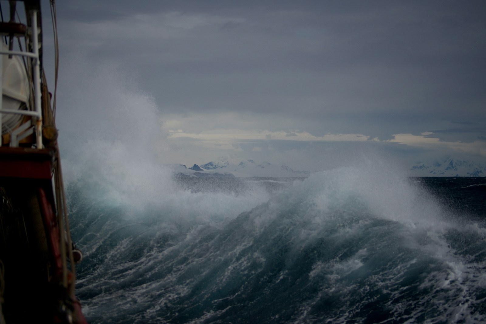 A tall wave breaking beside a ship's railing in a rough sea, with snow-covered ridges on the horizon