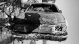 A heavily damaged car is positioned upside-down on a rocky and barren terrain. The car appears old with significant wear and tear, including missing parts and visible rust. Debris surrounds the vehicle, with rocks of various sizes scattered across the ground.
