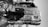 A heavily damaged car is positioned upside-down on a rocky and barren terrain. The car appears old with significant wear and tear, including missing parts and visible rust. Debris surrounds the vehicle, with rocks of various sizes scattered across the ground.