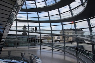Interior view of a modern, multi-level glass dome with spiral walkways and people walking on the paths. Exterior buildings and blue sky with clouds are visible through the glass.