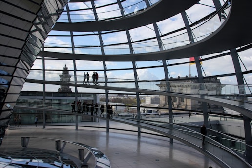 Interior view of a modern, multi-level glass dome with spiral walkways and people walking on the paths. Exterior buildings and blue sky with clouds are visible through the glass.