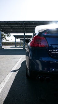A close-up of the rear end of a dark-colored car is visible, parked in a shaded area beneath a large metal awning. Sunlight creates a bright glow above, casting a shadow on the pavement. The car's design includes rounded taillights and dual exhaust pipes. In the background, the surrounding environment includes some trees and a clear sky.