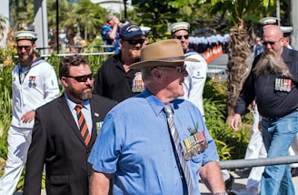 A group of men wearing various uniforms and medals are walking outdoors in a parade setting. Some are in military uniforms, including naval attire, while others are in suits with ties. The background includes greenery and a line of marching individuals.