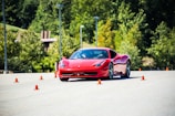 A sleek red sports car navigates through a series of orange traffic cones on a clear day, set against a backdrop of lush green trees. The scene suggests a driving course or performance test with the emphasis on speed and control.