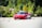 A sleek red sports car navigates through a series of orange traffic cones on a clear day, set against a backdrop of lush green trees. The scene suggests a driving course or performance test with the emphasis on speed and control.