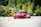 A sleek red sports car navigates through a series of orange traffic cones on a clear day, set against a backdrop of lush green trees. The scene suggests a driving course or performance test with the emphasis on speed and control.