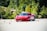 A sleek red sports car navigates through a series of orange traffic cones on a clear day, set against a backdrop of lush green trees. The scene suggests a driving course or performance test with the emphasis on speed and control.