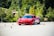 A sleek red sports car navigates through a series of orange traffic cones on a clear day, set against a backdrop of lush green trees. The scene suggests a driving course or performance test with the emphasis on speed and control.