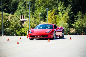 A sleek red sports car navigates through a series of orange traffic cones on a clear day, set against a backdrop of lush green trees. The scene suggests a driving course or performance test with the emphasis on speed and control.