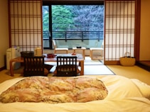 A traditional Japanese room featuring a futon on the tatami mat floor, with a low wooden table surrounded by floor chairs. Sliding shoji doors frame a large window, providing a view of a terrace with chairs and a backdrop of trees.