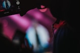 A person seen in silhouette holding a smartphone capturing a video. The background is blurred with vibrant colors of pink and blue, creating a dynamic atmosphere reminiscent of a live event or concert. The smartphone screen is clearly visible, showing a figure in a white outfit on stage.