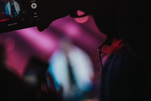 A person seen in silhouette holding a smartphone capturing a video. The background is blurred with vibrant colors of pink and blue, creating a dynamic atmosphere reminiscent of a live event or concert. The smartphone screen is clearly visible, showing a figure in a white outfit on stage.