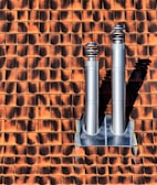Two metallic chimneys are mounted on a textured orange and brown tile roof. The tiles have a gradient shading effect with shadows casted by the chimneys.