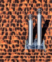 Two metallic chimneys are mounted on a textured orange and brown tile roof. The tiles have a gradient shading effect with shadows casted by the chimneys.