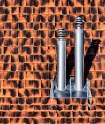 Two metallic chimneys are mounted on a textured orange and brown tile roof. The tiles have a gradient shading effect with shadows casted by the chimneys.