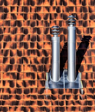 Two metallic chimneys are mounted on a textured orange and brown tile roof. The tiles have a gradient shading effect with shadows casted by the chimneys.