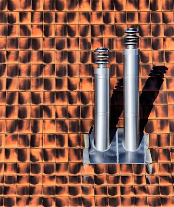 Two metallic chimneys are mounted on a textured orange and brown tile roof. The tiles have a gradient shading effect with shadows casted by the chimneys.