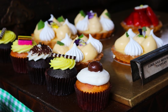 An assortment of colorful pastries is arranged on a wooden and brass surface. Several cupcakes with different toppings like chocolate shavings, cherries, and striped chocolate pieces are in the foreground. Lemon-flavored meringue tarts topped with white peaks and garnished with small pieces of green garnish and purple flowers are positioned behind the cupcakes. A tag labeled 'Lime and Meringue Tartlet' is visible.