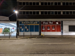 A dimly lit street scene featuring an empty commercial building with storefronts for 'Redfields' and 'Pizza Hut'. The surrounding area is deserted and there are visible street lamps, but no people are present.