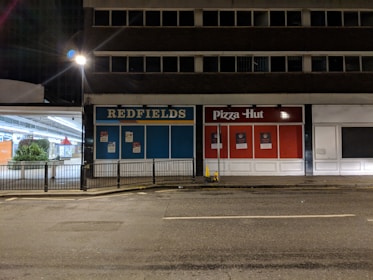 A dimly lit street scene featuring an empty commercial building with storefronts for 'Redfields' and 'Pizza Hut'. The surrounding area is deserted and there are visible street lamps, but no people are present.