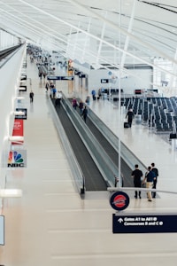 A spacious airport terminal with a long moving walkway runs through the center. Several people walk alongside or ride the walkway, carrying luggage or engaging in conversation. Overhead, the architecture features an intricate design of white beams and ample natural light streaming in. Various signs hang from the ceiling, directing travelers to gates A1 to A61, as well as Concourses B and C. The terminal is clean and modern, with empty seating areas to the right.