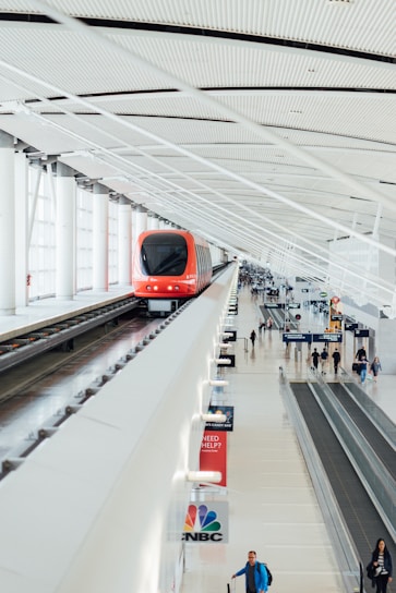 A modern, red automated train travels along an elevated track inside a spacious, well-lit terminal with a high, ribbed ceiling. Below, a few passengers walk or stand on moving sidewalks. Several signs, including one for CNBC, are visible among the terminal's clean lines and bright atmosphere.