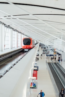 A modern, red automated train travels along an elevated track inside a spacious, well-lit terminal with a high, ribbed ceiling. Below, a few passengers walk or stand on moving sidewalks. Several signs, including one for CNBC, are visible among the terminal's clean lines and bright atmosphere.