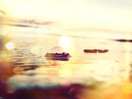 A tranquil seascape with sunlight reflecting off the water, creating a warm glow and bokeh effect. There are small ripples and bubbles on the water's surface, enhancing the serene ambiance.