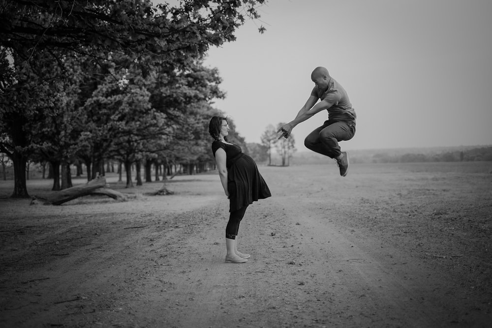 grayscale photo of man jumping in front of pregnant woman near trees during daytime