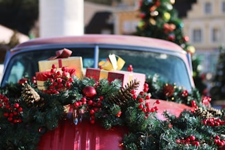 A vintage car is adorned with festive holiday decorations, including pine cones, red berries, and a lush green garland. Gifts wrapped in colorful paper with bows are piled on the car, while a Christmas tree can be seen in the background, adding to the celebratory atmosphere.