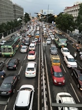 A densely packed urban traffic scene featuring a variety of vehicles, including buses, cars, and motorbikes, moving in both directions on a multi-lane road. Trees and buildings line the sides of the street, with traffic signals and streetlights visible. The atmosphere is busy and congested, typical of metropolitan rush hour.
